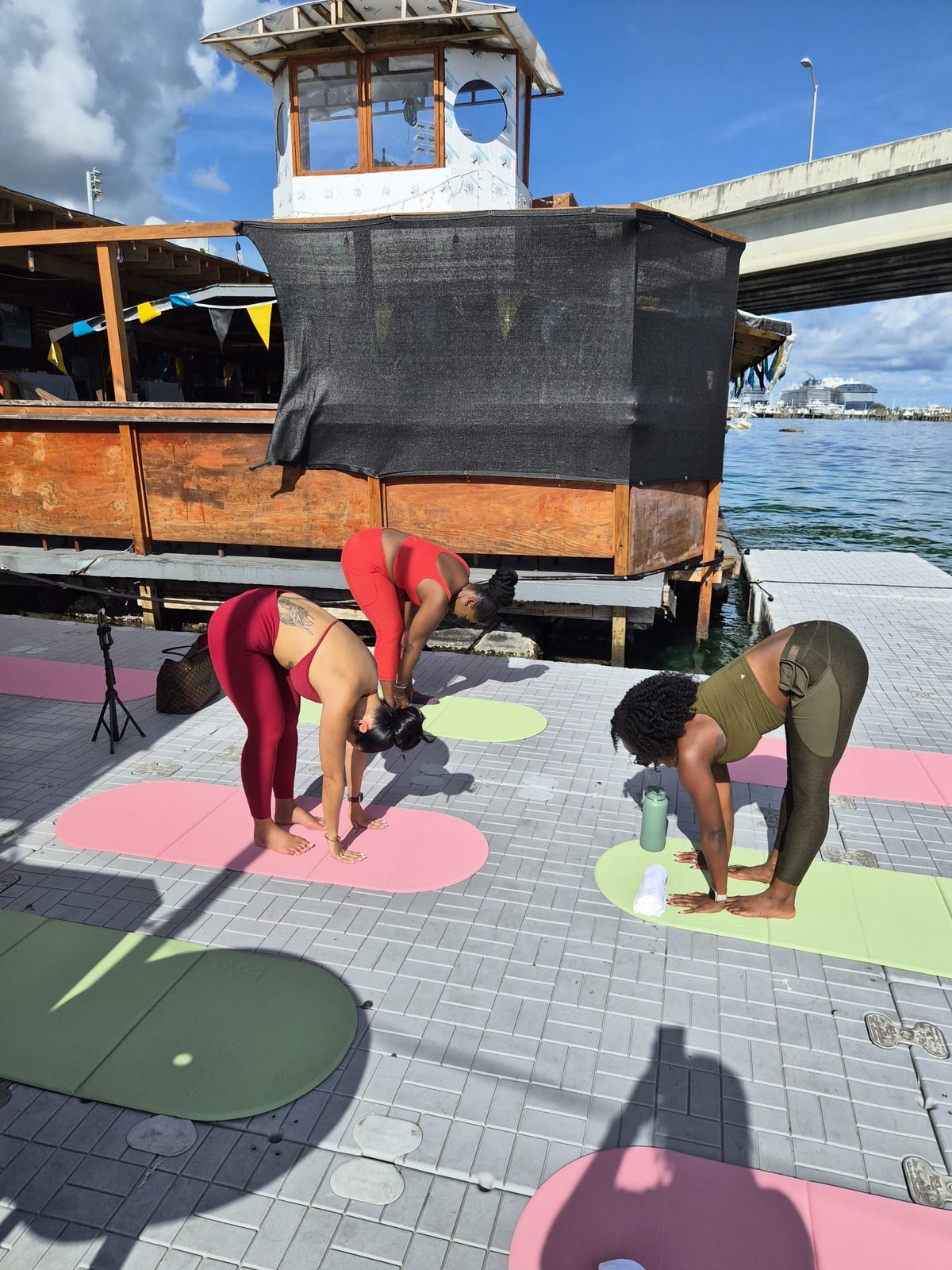 Two guests folding forward on pink and sage mats on a floating dock, with the Nassau bridge and harbour behind