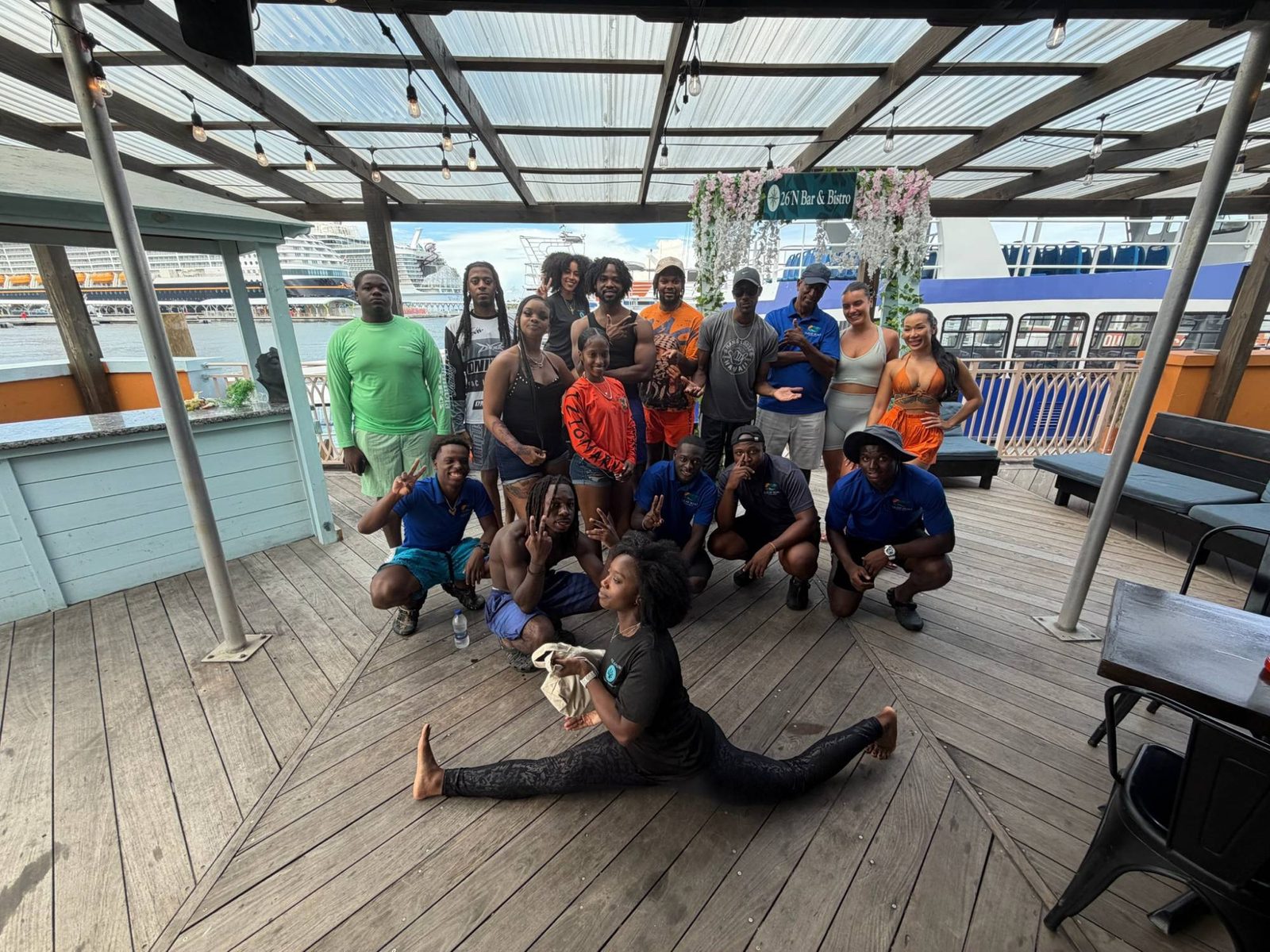 A group of smiling people posing together on a harbourside deck, with an instructor doing a split at the front