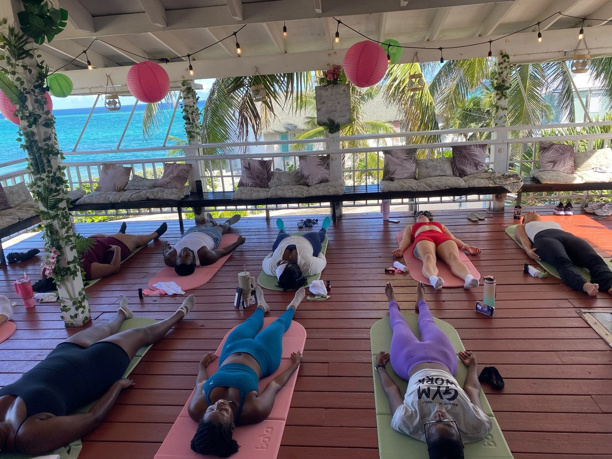 A Pilates brunch class lying on mats on a wooden sea-view deck under string lights