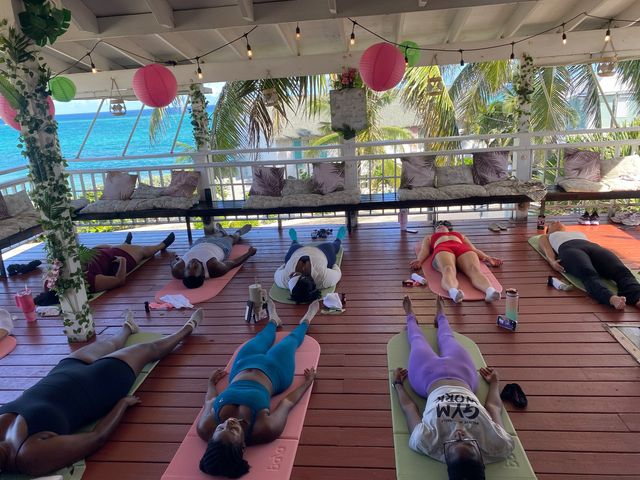 A Pilates brunch class lying on mats on a wooden sea-view deck under string lights