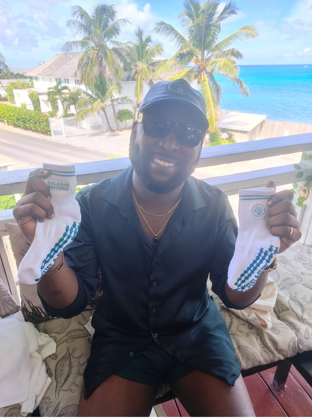 A smiling guest in a cap and sunglasses holding up two Fuel & Flow grip socks on a seaside terrace