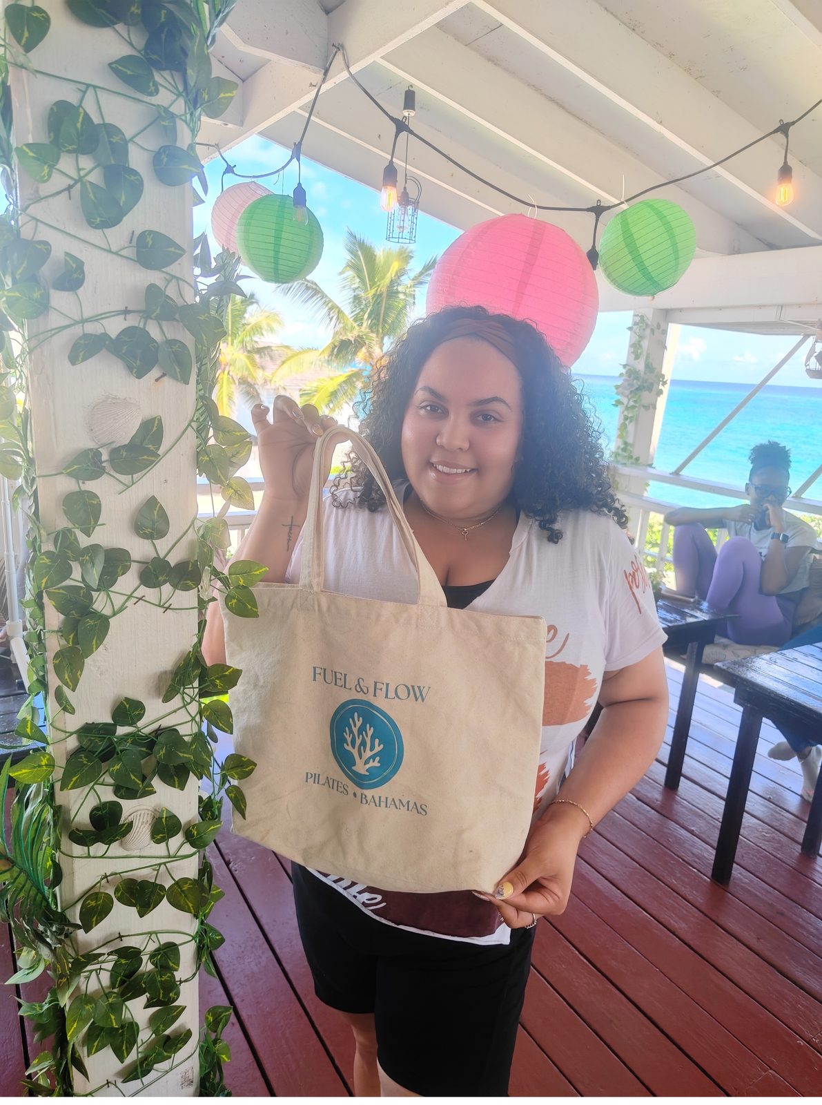 A guest holding a Fuel & Flow Pilates Bahamas canvas tote on a terrace with lanterns and sea behind